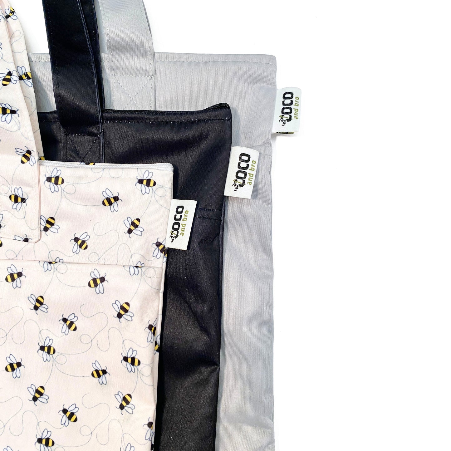 A collection of three zipped tote bags in black, grey and bumble bee designs, made from bamboo fibres in a bumble bee design, featuring two zipped sections and a handy loop tab to easily roll up the bag for storage. Image shows the bags zoomed in, with the Coco and Bro label visible.